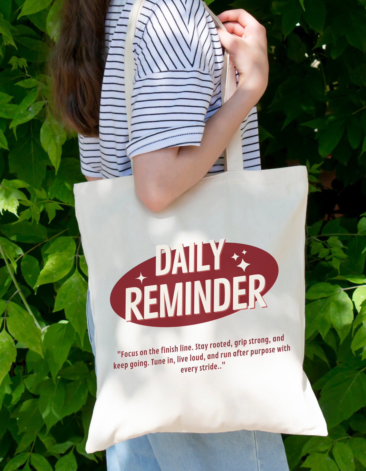 Person holding a tote bag with 'Daily Reminder' text against a green leafy background